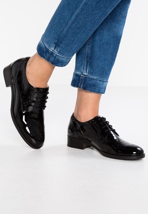 Chaussures brogues en cuir noir brillant avec lacets, portées avec un jean en denim bleu retroussé, sur une personne debout sur un sol blanc.