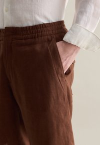 Pantaloni in lino marrone con vita elastica, tasche laterali e vestibilità rilassata. Il tessuto ha un aspetto testurizzato e una finitura opaca.