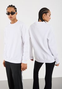 Two people with braided hair wear white long-sleeve tops; one faces forward with sunglasses, the other shows the back, both in black pants.