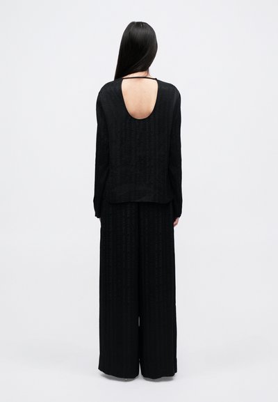 Svart jumpsuit med lange ermer, en åpen ryggdetalj, teksturert stoff med vertikale mønstre, og bred ben silhuett.