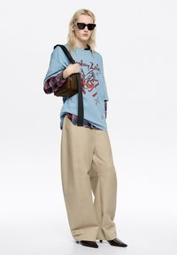 Camiseta gráfica azul con diseño rojo, superpuesta a una camisa de cuadros. Pantalones beige oversized y botas puntiagudas oscuras. Bolsa de hombro marrón y gafas de sol.