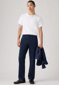 Jeune homme portant un t-shirt blanc, un pantalon bleu marine, des chaussures noires, tenant une veste en denim bleu marine dans la main droite, debout devant un fond uni.