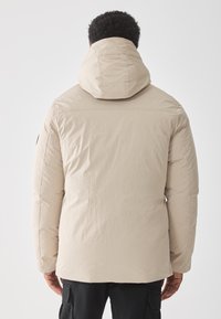 Giacca isolante beige con un grande cappuccio, texture trapuntata e taglio dritto. Presenta cuciture sottili e un logo sulla manica sinistra.
