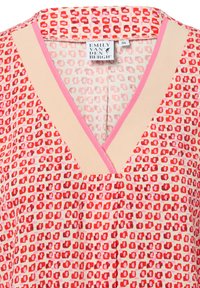 Czerwona bluzka w wzory z dekoltem w kształcie V. Posiada mieszankę geometrycznych kształtów w odcieniach czerwieni i różu. Materiał wydaje się lekki i gładki.