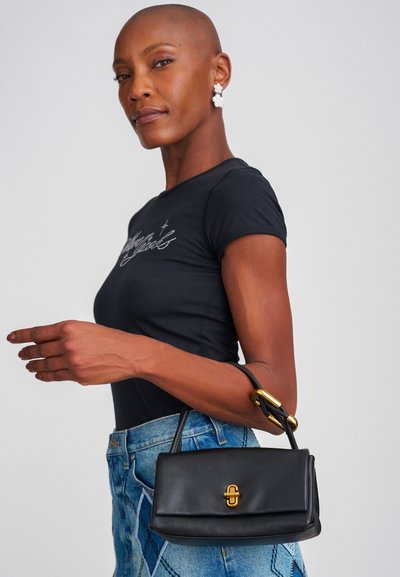 Bolso de mano de cuero negro con un diseño estructurado, cierre de hardware dorado y un asa corta. El modelo lleva una camiseta negra ajustada y unos vaqueros con patrones.