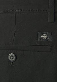 Black fabric pocket with a visible black button and a dark blue label featuring a logo, stitched details, and smooth texture.