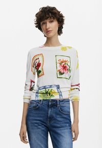 White sweater featuring colorful floral and label prints. Ribbed cuffs and hem. Paired with blue denim jeans.