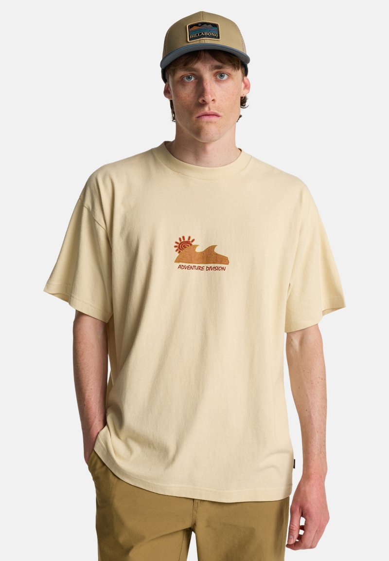 Μπεζ φαρδιά μπλούζα από βαμβάκι, με γραφικό σχέδιο σε γήινες αποχρώσεις που γράφει "ADVENTURE DIVISION" και σχέδιο κύματος.