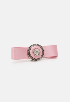 Ružičasta saten traka s okruglom srebrnom i ružičastom metalnom kopčom koja sadržava dekorativni uzorak, obrubljena geometrijskim detaljima.