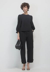 Schwarzes Oberteil mit Puffärmeln und subtilen Lochstickereien, kombiniert mit lockeren schwarzen Hosen mit Seitentaschen und umgeschlagenen Säumen.