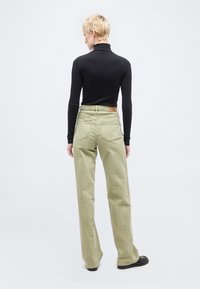 Άτομο με κοντά ξανθά μαλλιά, ντυμένο με μαύρο ζιβάγκο και φαρδιά ανοιχτό πράσινα παντελόνια, στέκεται στραμμένο προς ένα λευκό φόντο.