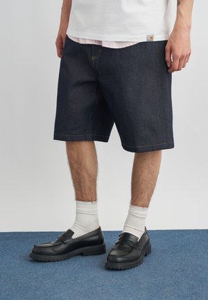 Hombre con pantalones cortos de mezclilla oscuros, calcetines blancos de tipo deportivo y mocasines de cuero negro, de pie sobre una alfombra azul.