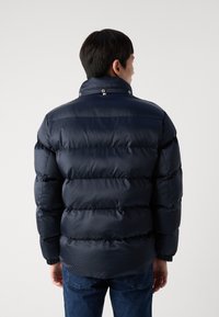 Marinblå pufferjacka med texturerad yta, rund krage och tryckknappsstängning. Har horisontella quiltade sektioner och ribbade manschetter.