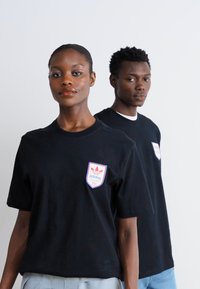 Melni kokvilnas t-krekli ar īsām piedurknēm, ar krāsainu Adidas logo uzlīmi uz krūtīm. Vienkāršs dizains un brīvs piegriezums.