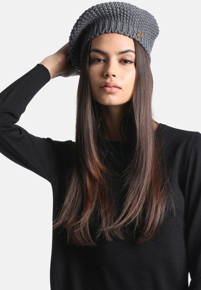 Femme aux longs cheveux bruns et raides, portant un pull noir et un béret gris texturé en tricot, tenant le béret d'une main.