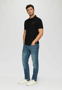 Schwarzes Polo-Shirt mit kurzen Ärmeln, kombiniert mit hellblauen Jeans und weißen Sneakers. Das Polo hat ein kleines goldenes Logo.