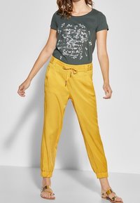 Grijze shirt met witte grafische print, gele joggingbroek met elastische boorden en platte sandalen. Glad materiaal en ontspannen pasvorm.