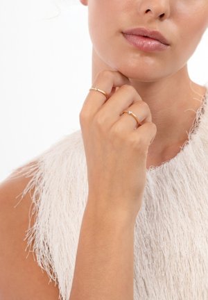 Two delicate gold rings on a hand, one featuring a small diamond accent. The person wears a textured white top with fringe details.