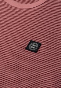 Mauvenvärinen waffle-neulottu kangas, jossa pyöreä pääntie ja pieni musta neliönmuotoinen logoplässi, jossa geometrinen merkki.