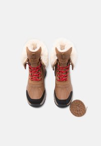 Bruine waterdichte laarzen met namaakbonten voering, rode veters, zwarte rubberen zolen en een ronde bruine veteraccessoire op een witte achtergrond.