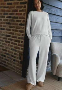 Weißes, strukturiertes Loungewear-Set mit Rippenmuster, bestehend aus einem langärmeligen Oberteil und Hose mit Kordelzug, kombiniert mit braunen Hausschuhen.