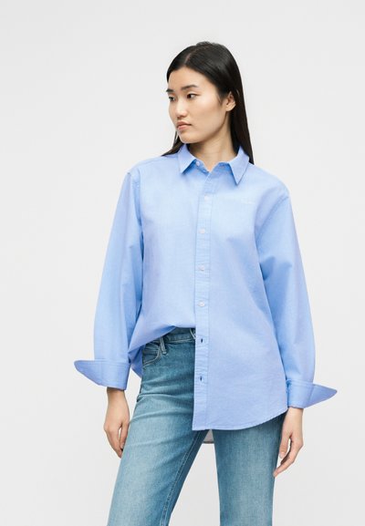 Νεαρή γυναίκα που φοράει μια άνετη ανοιχτό μπλε πουκάμισο με κουμπιά και μανίκια γυρισμένα, καθώς και τζιν μεσαίου τυπώματος, στέκεται μπροστά σε ένα απλό φόντο.