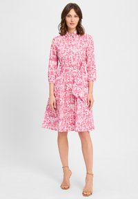 Vestito rosa con bottoni, vita aderente, maniche 3/4 e gonna svasata con un motivo tropicale. Cintura annodata in vita per un tocco in più.
