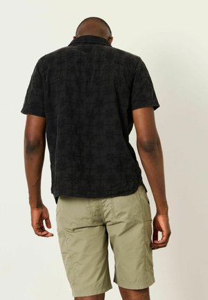 Man standing facing away wearing a black short-sleeve patterned shirt and light khaki cargo shorts against a plain background.