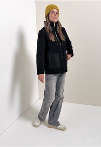 Giacca in pile nera con zip frontale, grande tasca nera, abbinata a jeans grigi e un berretto lavorato a maglia giallo. Sfondo neutro.