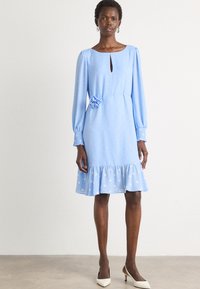 Lichtblauwe jurk met lange poefmouwen, stippenmotief, sleufhals, bloemaccent bij de taille en een rucheszoom. G gedragen met witte hakken.