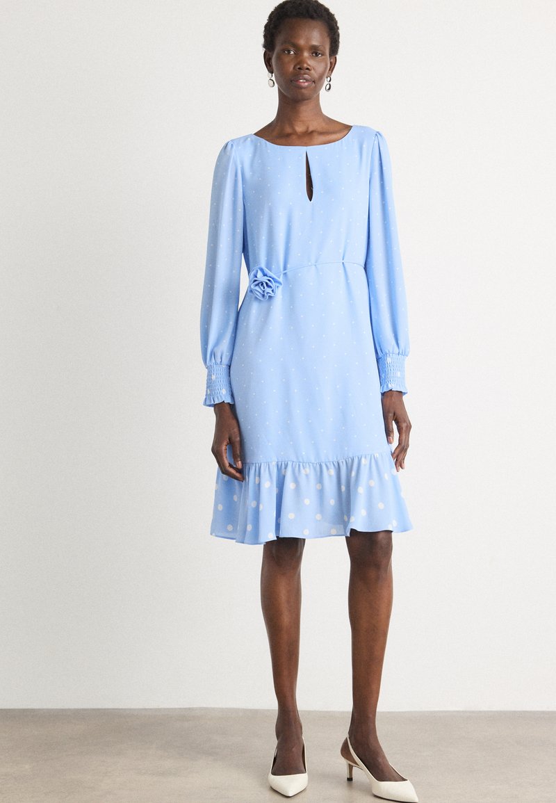 Lichtblauwe jurk met lange poefmouwen, stippenmotief, sleufhals, bloemaccent bij de taille en een rucheszoom. G gedragen met witte hakken.