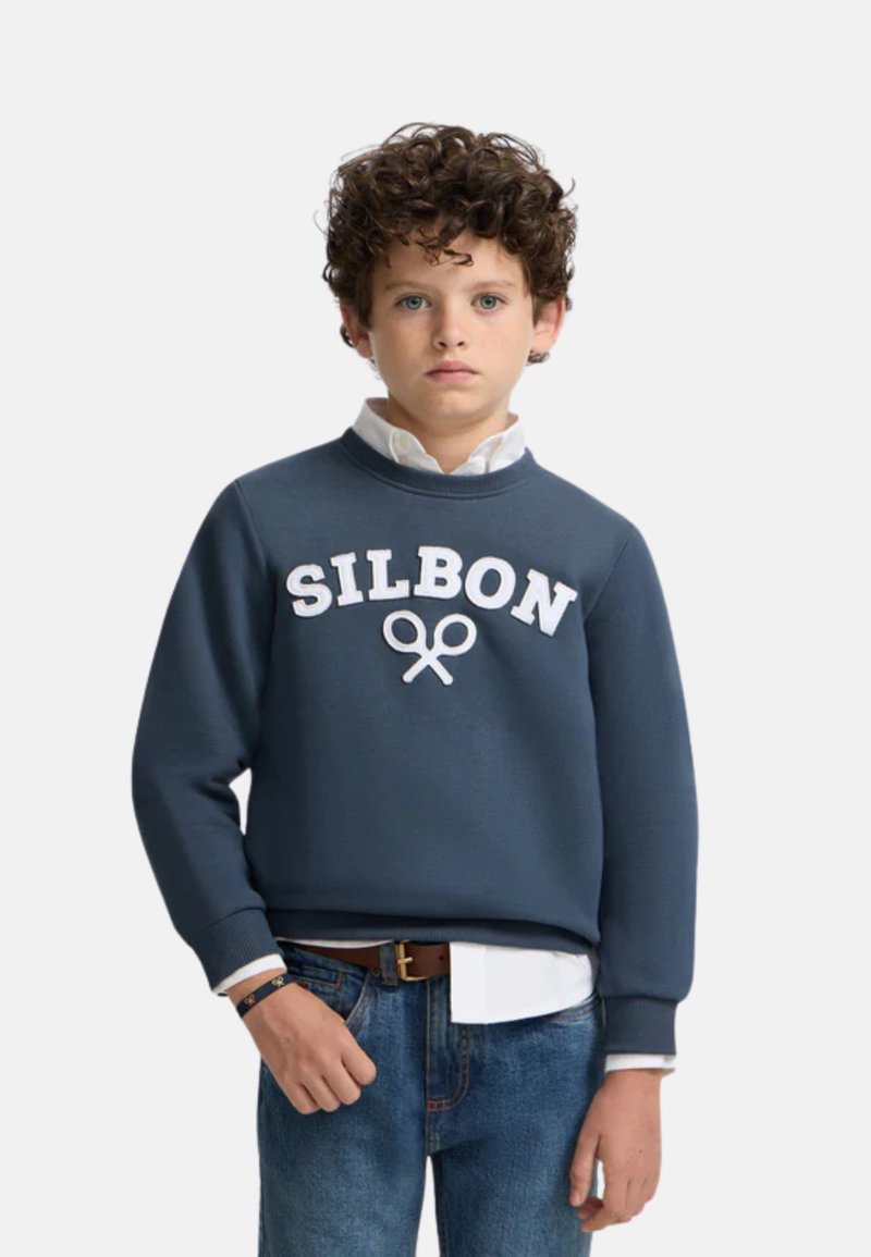 Sudadera navy con logotipo blanco "SILBON" y gráfico de tijeras. Cuello redondo y mangas largas. Lleva puesta sobre una camisa blanca con cuello y jeans de mezclilla.