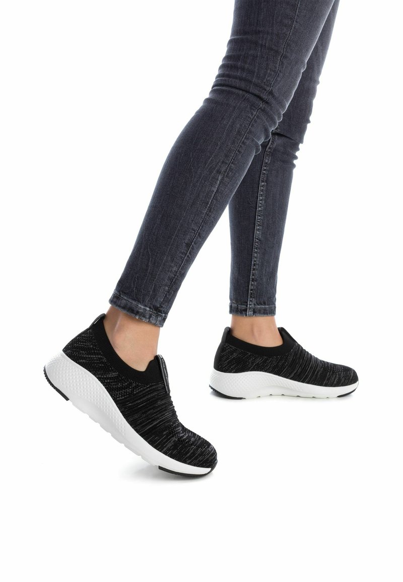 Zapatillas slip-on en blanco y negro con upper de punto texturizado, que presentan una suela gruesa y un cuello elástico, combinadas con jeans ajustados oscuros.