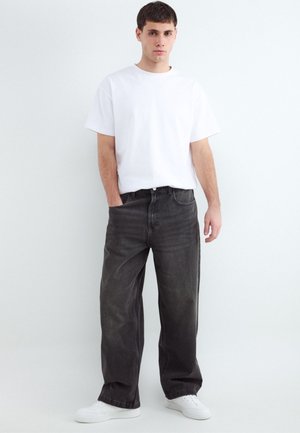 Young man standing wearing a plain white t-shirt, loose black jeans, and white sneakers against a white background.