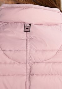 Vaate vaaleanpunainen toppaliivi, jossa on korkea kaulus, vinosti ommeltu kangas ja pieni musta etiketti logolla takana. Pehmeä ja sileä pinta.