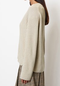 Maglione beige a coste con maniche lunghe, caratterizzato da una vestibilità rilassata e un motivo testurizzato. Indossato sopra una gonna a quadretti marrone.