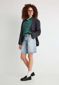 Νέα γυναίκα με σγουρά μαλλιά που φορά γυαλιά, πράσινη ριγέ πόλο μπλούζα, σκούρο σακάκι, τζιν σορτς, λευκές κάλτσες και μαύρα loafers, στέκεται μπροστά σε λευκό φόντο.