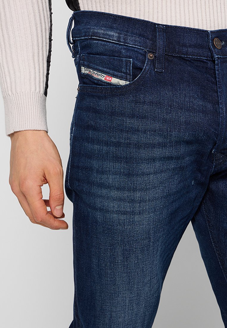 Homme portant un jean en denim bleu foncé et un pull côtelé beige clair, avec une petite étiquette de logo visible sur la poche avant.