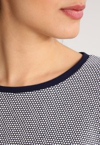 Marinblus med vitt mönster som har en strukturerad stickning, en vid halsringning och en ribbad krage. Närbild på tyg- och halsringningsdetaljer.