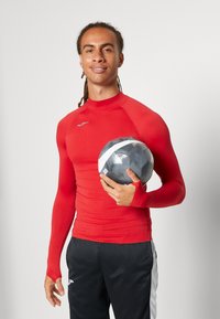 Röd långärmad träningsskjorta med tumhål, åtsittande design. Svarta byxor med vita detaljer. Modellen håller i en svart och grå fotboll.