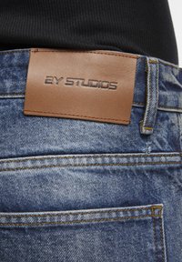 Étiquette en cuir marron avec l'inscription "2Y STUDIOS" embossée sur un jean en denim bleu porté avec un haut côtelé noir visible à la taille.