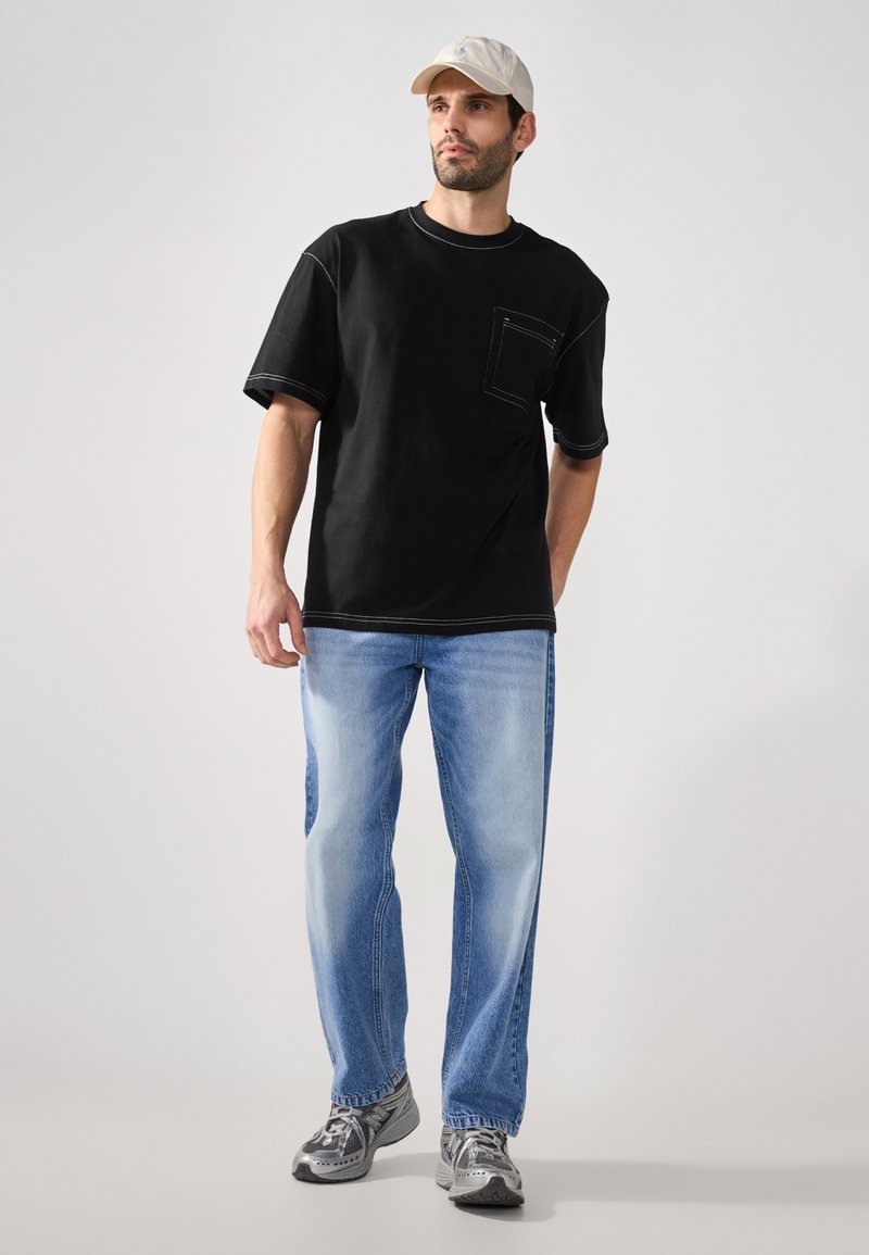 T-shirt noire à manches courtes avec une poche, associé à un jean en denim bleu clair. Complété par des chaussures de sport grises et une casquette beige.