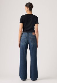 Γυναίκα που στέκεται δίνοντας την πλάτη της, φορώντας μαύρο μπλουζάκι με κοντά μανίκια και ψηλόμεσα, φαρδιά μπλε τζιν με πίσω τσέπες.