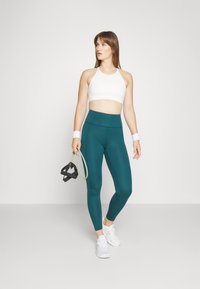 Tarkakék, magas derekú leggings és egy fehér sportmelltartó. A modell egy ellenállás szalagot tart. A fehér edzőcipők és a csuklópántok teszik teljessé az öltözetet.