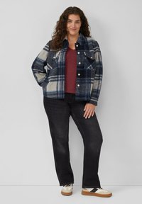 Karierte marineblaue und cremefarbene Flanelljacke über einem kastanienbraunen Oberteil, kombiniert mit schwarzen Jeans und beigen Sneakers mit schwarzen Akzenten, vor einem hellen Hintergrund.