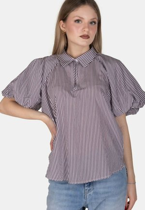 Giovane donna con capelli lisci che indossa una blusa a righe a maniche corte con collo e jeans azzurri, posando con una mano sui fianchi.