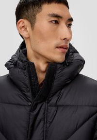 Chaqueta acolchada negra con cuello alto y capucha ajustable, con botones a presión y un diseño acolchado. Textura suave, apariencia ligera.