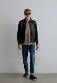 Fekete bőrdzseki bézs ing fölött, kék farmerrel és fekete cipővel párosítva. A modell napszemüveget visel, semleges háttér előtt áll.