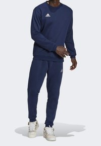 Sweat-shirt bleu marine et pantalon de jogging assorti en tissu doux. Présente un logo Adidas blanc sur la poitrine et le pantalon. Des baskets blanches complètent la tenue.