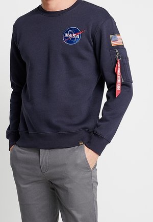 Sudadera azul marino con el logo de NASA, cuello acanalado y bolsillo en la manga con un llavero rojo. Combinando con pantalones grises de corte relajado.